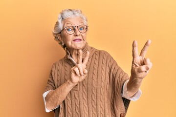 Senior grey-haired woman wearing casual clothes and glasses smiling looking to the camera showing fingers doing victory sign. number two.