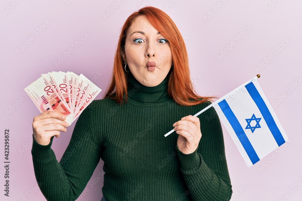 Poster beautiful redhead woman holding 20 shekels banknotes and israel flag making fish face with mouth and