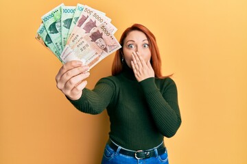 Beautiful redhead woman holding south korean won banknotes covering mouth with hand, shocked and afraid for mistake. surprised expression