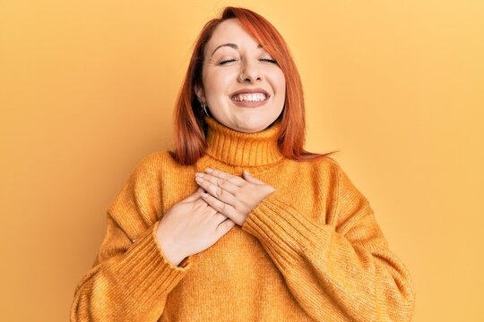 Beautiful redhead woman wearing casual winter sweater over yellow background smiling with hands on chest with closed eyes and grateful gesture on face. health concept.