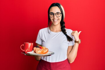 Young hispanic woman wearing waitress apron holding tray with breakfast pointing thumb up to the side smiling happy with open mouth