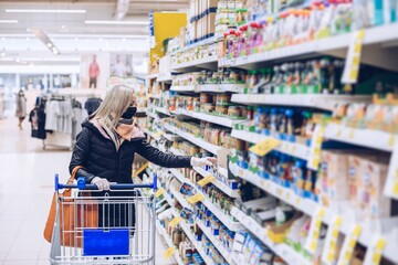 Woman in a protectivel mask and rubber gloves stands in a supermarket with a shopping cart and watchs products.
