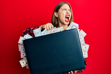 Beautiful young blonde woman holding briefcase full of dollars angry and mad screaming frustrated and furious, shouting with anger looking up.