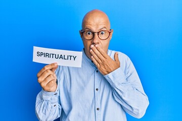 Middle age bald man holding paper with spirituality word covering mouth with hand, shocked and...