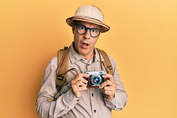 Middle age bald man wearing explorer hat and vintage camera in shock face, looking skeptical and sarcastic, surprised with open mouth
