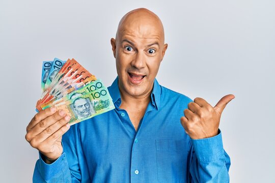 Middle age bald man holding australian dollars pointing thumb up to the side smiling happy with open mouth