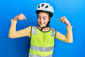 Beautiful brunette little girl wearing bike helmet and reflective vest showing arms muscles smiling proud. fitness concept.