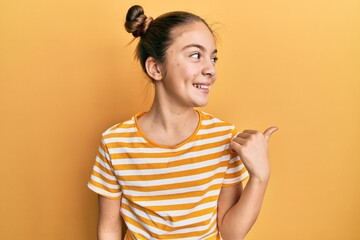 Beautiful brunette little girl wearing casual striped t shirt smiling with happy face looking and pointing to the side with thumb up.