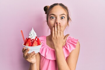 Beautiful brunette little girl eating strawberry ice cream covering mouth with hand, shocked and...
