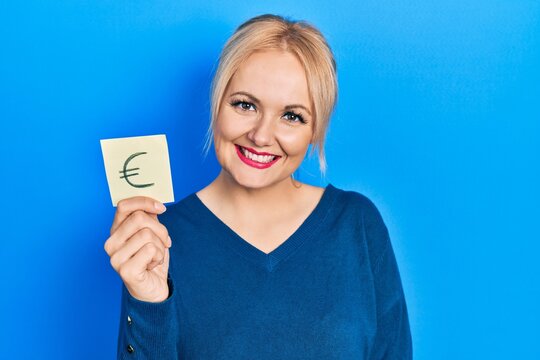 Young blonde woman holding euro symbol reminder looking positive and happy standing and smiling with a confident smile showing teeth