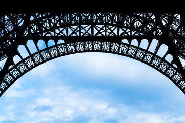 Arched detail of the Eiffel Tower silhouetted against a cloudy blue sky.