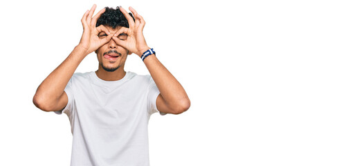 Young arab man wearing casual white t shirt doing ok gesture like binoculars sticking tongue out, eyes looking through fingers. crazy expression.