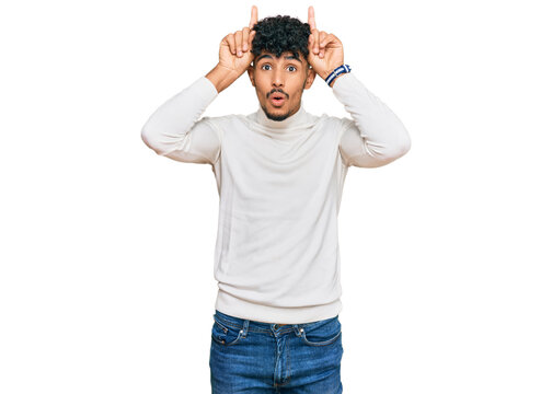 Young arab man wearing casual winter sweater doing funny gesture with finger over head as bull horns