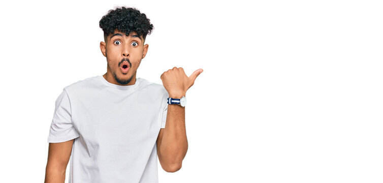 Young arab man wearing casual white t shirt surprised pointing with hand finger to the side, open mouth amazed expression.