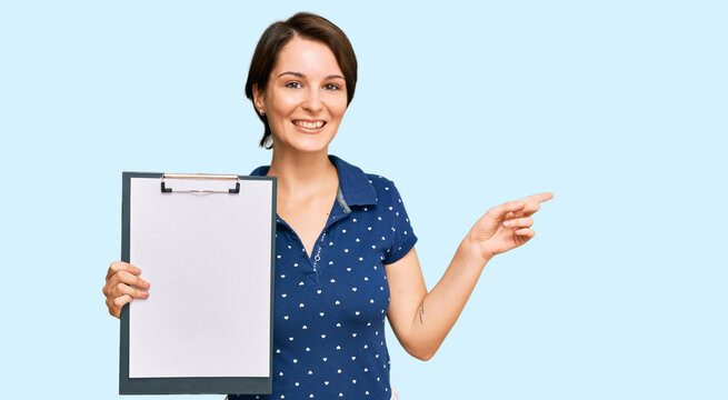 Young brunette woman with short hair holding clipboard with blank space smiling happy pointing with hand and finger to the side