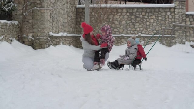 vacation, game, family concept - slo-mo authentic three happy preschool toddler kids siblings with young wonan mom sledding and play with snow. snowfall in cold season weather in winter yard outdoors