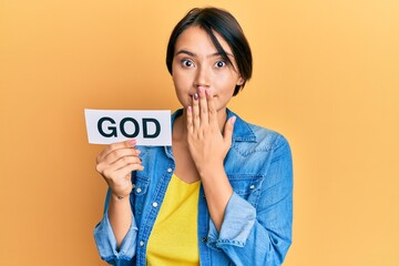 Beautiful young woman with short hair holding paper with god word covering mouth with hand, shocked...