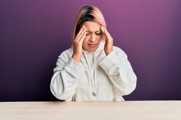 Hispanic woman with pink hair wearing casual sweatshirt sitting on the table with hand on head for...