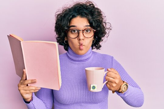 Young hispanic woman with curly hair reading a book and drinking a cup of coffee making fish face with mouth and squinting eyes, crazy and comical.