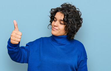 Young hispanic woman with curly hair wearing turtleneck sweater looking proud, smiling doing thumbs up gesture to the side