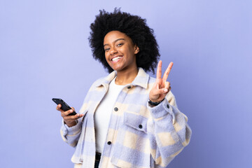 Young African American woman using mobile phone isolated on purple background smiling and showing victory sign