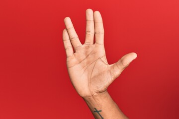 Hand of hispanic man over red isolated background greeting doing vulcan salute, showing hand palm and fingers, freak culture - obrazy, fototapety, plakaty