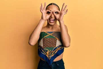 Hispanic man wearing make up and long hair wearing womans top doing ok gesture like binoculars sticking tongue out, eyes looking through fingers. crazy expression.