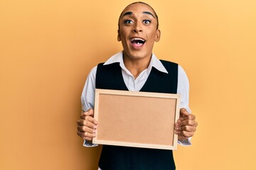 Hispanic man wearing make up and long hair holding empty corkboard celebrating crazy and amazed for success with open eyes screaming excited.