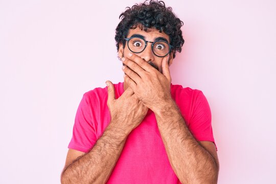 Handsome young man with curly hair and bear wearing casual clothes and glasses shocked covering mouth with hands for mistake. secret concept.