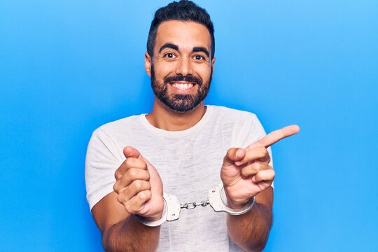 Young hispanic man wearing prisoner handcuffs smiling happy pointing with hand and finger to the side