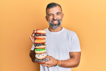 Middle age handsome man holding tasty colorful doughnuts winking looking at the camera with sexy...