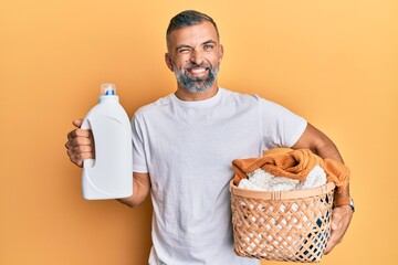 Middle age handsome man holding laundry basket and detergent bottle winking looking at the camera...