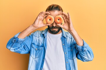 Attractive man with long hair and beard holding tasty colorful doughnuts on eyes skeptic and...