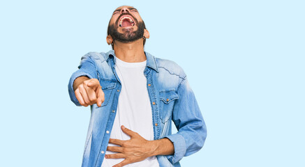 Attractive man with long hair and beard wearing casual denim jacket laughing at you, pointing finger to the camera with hand over body, shame expression