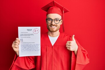 Young redhead man wearing graduation robe showing passed exam smiling happy and positive, thumb up...