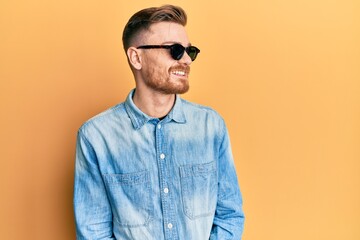 Young redhead man wearing stylish sunglasses looking away to side with smile on face, natural expression. laughing confident.