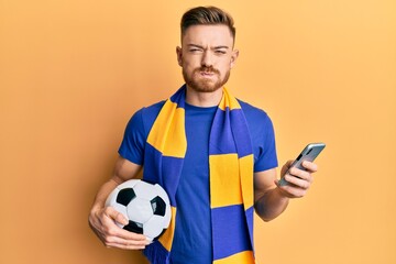 Young redhead man wearing hooligan scarf and using smartphone puffing cheeks with funny face. mouth inflated with air, catching air.