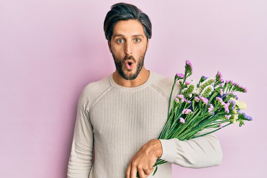 Young hispanic man holding flowers scared and amazed with open mouth for surprise, disbelief face