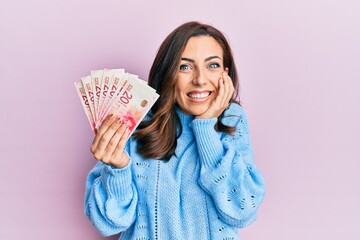 Young brunette woman holding 20 israel shekels banknotes laughing and embarrassed giggle covering mouth with hands, gossip and scandal concept