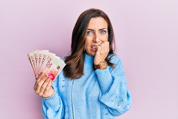 Young brunette woman holding 20 israel shekels banknotes looking stressed and nervous with hands on mouth biting nails. anxiety problem.