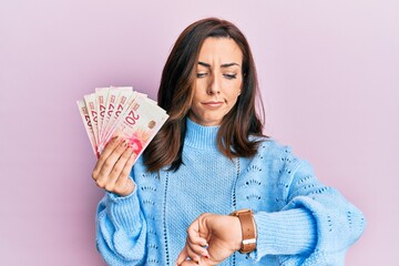 Young brunette woman holding 20 israel shekels banknotes checking the time on wrist watch, relaxed and confident
