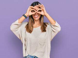 Young woman wearing casual clothes and glasses doing ok gesture like binoculars sticking tongue out, eyes looking through fingers. crazy expression.
