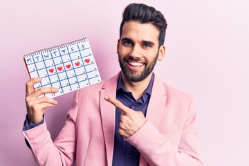 Young handsome man with beard holding calendar with hearts smiling happy pointing with hand and finger