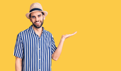 Young handsome man with beard wearing summer hat and striped shirt smiling cheerful presenting and pointing with palm of hand looking at the camera.