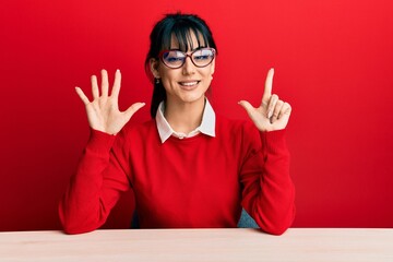 Young brunette woman with bangs wearing glasses sitting on the table showing and pointing up with...