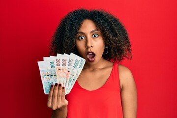 Beautiful african american woman with afro hair holding czech koruna banknotes scared and amazed with open mouth for surprise, disbelief face