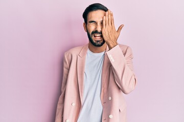 Young hispanic man wearing business jacket covering one eye with hand, confident smile on face and surprise emotion.