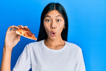 Young chinese woman eating tasty pepperoni pizza scared and amazed with open mouth for surprise, disbelief face