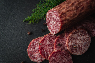 Sliced salami sausage on a board. Meat product and herbs on a dark background. A quick meal or snack. Dried sausage with black peppercorns.