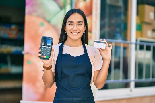 Young latin shopkeeper girl smiling happy holding credit card and dataphone at fruit store.